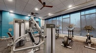 Located at 434 W Lake St in Minneapolis, MN, this fitness center offers a thoughtfully designed, single-floor exercise space ideal for a comprehensive workout experience. The room is finished with a drop tile ceiling equipped with recessed lighting and ceiling fans, maintaining a bright and comfortable environment throughout. Bold teal accent walls contrast with clean white surfaces, giving the space a modern and energetic aesthetic. Flooring transitions between durable carpet tile and rubberized mat sections, each suited to the type of activity performed in that zone. A large bank of floor-to-ceiling windows lines one wall, fitted with roller shades to manage light and glare while still allowing views of the surrounding Minneapolis streetscape. Full-length mirrors are mounted across a dedicated strength-training wall, visually expanding the space and supporting proper form during exercise. The room is outfitted with a diverse selection of Life Fitness cardio equipment, including treadmills, ellipticals, and stationary bikes, as well as a multi-functional cable strength machine and additional resistance equipment. A small workstation with a desk and shelving is tucked into one corner, providing a practical administrative or check-in area. The overall layout is organized and efficient, maximizing utility within the available footprint.