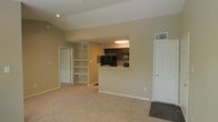 This single-floor space is designed with an open-concept layout that promotes a sense of spaciousness and functional flow. The living area is finished with wall-to-wall neutral carpeting and painted in warm beige tones, creating a welcoming and versatile environment.

A standout feature of the space is the pass-through kitchen window, which connects the kitchen to the main living area, enhancing the open feel. The kitchen is equipped with dark espresso cabinetry, granite-style countertops, stainless steel appliances including a refrigerator and microwave, and a black range. Recessed lighting illuminates the kitchen, complementing the overall brightness of the space.

Adjacent to the living area, a set of built-in open shelves provides practical storage and display options. Recessed ceiling lights in the living room ensure consistent lighting throughout. Two interior doors are visible — one leading to an additional room and another providing access to a closet or utility space. A ceiling-mounted HVAC vent and wall-mounted thermostat reflect thoughtful climate control integration throughout the unit.