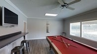 This single-floor recreational space is thoughtfully designed for entertainment and leisure. The centerpiece of the room is a full-size pool table with a bold red felt surface, complete with cues mounted on a wall-mounted rack nearby, making it immediately ready for use. The room features a combination of natural and artificial lighting, with a skylight panel in the ceiling flooding the space with daylight, complemented by a ceiling fan that ensures comfortable air circulation throughout. A window along one wall adds additional natural light while providing a connection to the outdoors. The flooring consists of durable wood-look planks in a warm grey-brown tone, offering both practicality and aesthetic appeal. Crown molding runs along the perimeter where the walls meet the ceiling, adding a polished, finished look to the space. A bar-height countertop with industrial-style metal stools lines one side of the room, creating a casual seating and refreshment area ideal for spectators or socializing. The walls are painted in a clean, neutral grey that gives the room a modern and cohesive feel. Overall, the space delivers a well-appointed, functional environment suited for hosting gatherings and recreational activities.