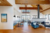 This single-story community amenity space combines striking architectural design with thoughtfully curated interiors. The centerpiece of the space is a soaring vaulted great room with exposed wooden beam ceilings and tongue-and-groove wood paneling overhead, creating a warm yet airy atmosphere. Crisp white walls contrast beautifully with the natural wood tones, while large black-framed glass doors and clerestory windows flood the space with abundant natural light and provide views of the surrounding community.

A bold, tile-clad freestanding fireplace with a cylindrical metal flue serves as the focal point of the lounge area, which is furnished with plush velvet armchairs, leather seating, and coffee tables arranged on patterned area rugs. A built-in banquette dining nook with pendant lighting and round bistro tables offers a casual dining setting along one wall. Decorative pendant lights and a geometric lantern chandelier add curated elegance throughout.

The fitness center is well-appointed across two areas: one featuring treadmills and cardio equipment positioned in front of a full-width glass garage door for an open, light-filled workout experience, and a second strength-training room with free weights, resistance machines, a rowing machine, exercise bike, and a warm wood accent wall. Carpet tile flooring and mirrored walls complete the practical yet polished fitness environment.