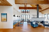 This single-story community amenity space combines striking architectural design with thoughtfully curated interiors. The centerpiece of the space is a soaring vaulted great room with exposed wooden beam ceilings and tongue-and-groove wood paneling overhead, creating a warm yet airy atmosphere. Crisp white walls contrast beautifully with the natural wood tones, while large black-framed glass doors and clerestory windows flood the space with abundant natural light and provide views of the surrounding community.

A bold, tile-clad freestanding fireplace with a cylindrical metal flue serves as the focal point of the lounge area, which is furnished with plush velvet armchairs, leather seating, and coffee tables arranged on patterned area rugs. A built-in banquette dining nook with pendant lighting and round bistro tables offers a casual dining setting along one wall. Decorative pendant lights and a geometric lantern chandelier add curated elegance throughout.

The fitness center is well-appointed across two areas: one featuring treadmills and cardio equipment positioned in front of a full-width glass garage door for an open, light-filled workout experience, and a second strength-training room with free weights, resistance machines, a rowing machine, exercise bike, and a warm wood accent wall. Carpet tile flooring and mirrored walls complete the practical yet polished fitness environment.