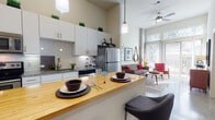 This single-floor residence showcases a thoughtfully designed open-concept layout that seamlessly connects the kitchen, dining, and living areas. The kitchen is a standout feature, boasting white cabinetry that extends to dramatic heights, a classic subway tile backsplash, granite countertops, stainless steel appliances, and a butcher block island with seating — ideal for casual dining and entertaining. Polished concrete floors run through the main living area, adding an industrial-chic aesthetic that complements the soaring ceilings and exposed concrete columns.

The living room is bright and airy, benefiting from floor-to-ceiling windows and a sliding glass door that opens to an outdoor space, flooding the interior with abundant natural light. A ceiling fan enhances comfort throughout the open area.

The bedroom offers a calm retreat with plush carpeting, a generous window framed by layered curtains and blinds, and a well-sized closet. The bathroom is clean and functional, featuring white cabinetry, granite countertops, polished concrete flooring, and a full-size tiled tub-shower combination. Overall, the space delivers a contemporary urban atmosphere with warm mid-century modern accents throughout.