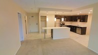 This single-floor residence offers a thoughtfully designed open-concept layout that seamlessly connects the living area and kitchen. The kitchen is a standout feature, showcasing rich dark cabinetry that contrasts beautifully with light countertops and a white kitchen island with bar-height seating — ideal for casual dining or entertaining. Stainless steel appliances, including a refrigerator, microwave, and range, add a modern, polished finish. Pendant lighting hangs above the island, providing warm, focused illumination that enhances the kitchen's contemporary aesthetic. Recessed lighting throughout ensures the space remains bright and inviting. The living area is finished with plush, neutral-toned carpeting that adds warmth and comfort underfoot. A coat closet near the entryway provides convenient storage upon arrival, while additional built-in shelving offers further organizational space. The unit's entry door is clearly visible from the main living area, contributing to a sense of openness and easy flow throughout the floor plan. The overall ambiance is clean, modern, and move-in ready, with a neutral color palette that provides a versatile backdrop for a variety of interior styles.