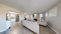 Located at 5272 Tamarus St in Las Vegas, NV, this single-floor unit offers a thoughtfully arranged layout encompassing a living room, dining room, and kitchen. The living area is anchored by a fireplace with a shiplap-style surround, framed by multiple windows that bring in natural light and create an open, airy atmosphere. Light wood-style flooring runs throughout, complementing the soft, neutral gray wall tones. An elegant arched passageway connects the living space to the kitchen, adding a distinctive architectural touch. The kitchen is outfitted with crisp white cabinetry, a classic white subway tile backsplash, gray countertops, and stainless steel appliances, including a full-size refrigerator and range. The dining area, positioned adjacent to the kitchen, features a window with slatted blinds and offers an intimate setting for meals. A textured ceiling adds visual character throughout the space. The overall design blends modern finishes with warm, welcoming details, resulting in a cohesive and functional living environment well-suited for comfortable everyday use in the heart of Las Vegas.