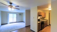 This single-floor residence offers a practical and comfortable layout designed for everyday living. The open floor plan creates a seamless flow between the kitchen and the main living area, making the space feel connected and easy to navigate.

The kitchen is outfitted with warm wood-toned cabinetry providing ample storage, a tile backsplash, granite-look countertops, and black appliances including a range. A range hood is mounted above the stove, and the space is efficiently arranged to maximize functionality.

The living area features plush carpeting, light-toned walls, and a ceiling fan with an integrated light fixture, contributing to a relaxed and airy atmosphere. A wall-mounted air conditioning unit ensures year-round comfort. The room is brightened by a sliding glass door that opens to a private balcony, allowing natural light to fill the space and offering a pleasant outdoor retreat.

The transition between the kitchen and living room is defined by durable hardwood-style flooring, adding visual contrast and easy maintenance near the entryway. Clean white walls throughout create a neutral backdrop that suits a variety of décor styles. Overall, the space is designed with practicality and comfort in mind, offering a well-organized and inviting environment.