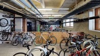 This bike storage room offers a practical and well-organized space designed to accommodate a large number of bicycles. The room features an industrial character, with exposed ceiling beams, overhead pipes, and fluorescent strip lighting that illuminate the concrete floors below. Multi-pane windows along one wall allow natural light to filter into the space, adding brightness to the utilitarian environment.

Bicycle parking is arranged throughout the room in multiple configurations. Wall-mounted vertical racks provide space-efficient storage along one section, while open floor areas accommodate additional bikes parked side by side. A plywood partition divides part of the room, creating distinct zones within the storage area. A green bike repair stand is also present, offering a convenient maintenance resource for cyclists.

A row of tall black metal lockers lines one wall, providing secure storage for helmets, gear, and personal belongings. The exposed brick and wood structural columns throughout add a raw, industrial character to the space. A colorful graffiti mural decorates one wall, lending a vibrant and urban artistic element to the room. A standard door provides direct access to the adjoining building interior, making this a highly functional and accessible amenity.