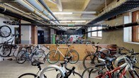 This bike storage room offers a practical and well-organized space designed to accommodate a large number of bicycles. The room features an industrial character, with exposed ceiling beams, overhead pipes, and fluorescent strip lighting that illuminate the concrete floors below. Multi-pane windows along one wall allow natural light to filter into the space, adding brightness to the utilitarian environment.

Bicycle parking is arranged throughout the room in multiple configurations. Wall-mounted vertical racks provide space-efficient storage along one section, while open floor areas accommodate additional bikes parked side by side. A plywood partition divides part of the room, creating distinct zones within the storage area. A green bike repair stand is also present, offering a convenient maintenance resource for cyclists.

A row of tall black metal lockers lines one wall, providing secure storage for helmets, gear, and personal belongings. The exposed brick and wood structural columns throughout add a raw, industrial character to the space. A colorful graffiti mural decorates one wall, lending a vibrant and urban artistic element to the room. A standard door provides direct access to the adjoining building interior, making this a highly functional and accessible amenity.