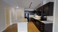This single-floor residence showcases a thoughtfully designed open-concept layout that seamlessly integrates living and kitchen areas. The kitchen is a standout feature, outfitted with rich, dark espresso cabinetry that extends to the ceiling, providing generous storage space. A central island with a clean, light-toned countertop offers additional prep space and serves as a natural gathering point. The kitchen is fully equipped with black appliances, including a refrigerator, range, built-in microwave, and dishwasher, all set against a subtle tile backsplash that adds texture to the space. Track lighting mounted on the ceiling illuminates the kitchen effectively, complementing the recessed lighting throughout. Warm hardwood-style flooring runs consistently across the space, adding a welcoming and cohesive aesthetic. The open entryway, featuring a light wood-toned door, provides a clean and modern transition into the home. The overall ambiance is contemporary and functional, with a neutral color palette of whites and dark browns that creates a balanced, modern feel. The layout promotes an easy flow between spaces, making it a practical and stylish residence.