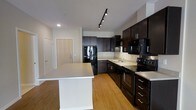 This single-floor residence showcases a thoughtfully designed open-concept layout that seamlessly integrates living and kitchen areas. The kitchen is a standout feature, outfitted with rich, dark espresso cabinetry that extends to the ceiling, providing generous storage space. A central island with a clean, light-toned countertop offers additional prep space and serves as a natural gathering point. The kitchen is fully equipped with black appliances, including a refrigerator, range, built-in microwave, and dishwasher, all set against a subtle tile backsplash that adds texture to the space. Track lighting mounted on the ceiling illuminates the kitchen effectively, complementing the recessed lighting throughout. Warm hardwood-style flooring runs consistently across the space, adding a welcoming and cohesive aesthetic. The open entryway, featuring a light wood-toned door, provides a clean and modern transition into the home. The overall ambiance is contemporary and functional, with a neutral color palette of whites and dark browns that creates a balanced, modern feel. The layout promotes an easy flow between spaces, making it a practical and stylish residence.