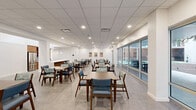 Located at 1320 Art Dilley Dr in Austin, TX, this single-floor community dining space offers a generous, open-plan layout designed to accommodate a variety of group settings. The room is furnished with multiple wooden dining tables and chairs upholstered in blue and patterned fabric, arranged throughout to encourage both intimate gatherings and larger social interaction.

A centrally positioned kitchenette serves as a functional anchor point, featuring warm wood cabinetry, a tiled backsplash in blue, pendant lighting, a stainless steel refrigerator, and a microwave — well-suited for food service or catering support. The ceiling is finished with a suspended acoustic tile grid outfitted with recessed lighting, providing consistent, even illumination throughout.

Large floor-to-ceiling windows and glass sliding doors line one side of the space, flooding the interior with natural light and offering direct views of and access to an outdoor courtyard area. The light-toned flooring features a subtle herringbone-style pattern, complementing the clean white walls. Colorful framed artwork adds visual interest and warmth to the otherwise minimal aesthetic. A wide corridor connects the dining area to the exterior, reinforcing the seamless indoor-outdoor flow of this thoughtfully designed communal space.