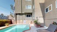 This single-floor unit is part of a multi-story residential building with a stucco exterior finish. One of its standout features is the covered balcony, which offers a direct view of the community pool area below, surrounded by palm trees and tropical landscaping that create a resort-like atmosphere. The balcony is finished with a smooth concrete floor and is enclosed by horizontal metal railings, providing an open yet secure outdoor space.

The balcony is furnished with a glass-top dining table and ornate wrought-iron chairs with striped cushioned seats, making it well-suited for outdoor dining or relaxing. A sliding glass door connects the balcony to the interior, allowing for easy indoor-outdoor flow and natural light. Steps on the balcony lead up to a door, suggesting additional access points within the space.

A no-smoking sign is posted on the balcony wall, indicating a smoke-free building policy. The building's exterior features textured stucco walls and multi-pane windows. The communal pool area below is well-maintained, flanked by lounge seating and lush greenery, adding to the overall appeal of the outdoor environment.
