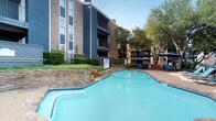 This outdoor amenity space in Dallas showcases a generously sized freeform swimming pool as its centerpiece, finished with teal-blue water and tiled edges that reflect the Texas sky above. A distinctive stacked-stone retaining wall adds a naturalistic, rustic charm to the pool's edge, contrasting beautifully with the clean, curved lines of the pool itself. The surrounding deck provides ample space for lounging, furnished with blue chaise lounge chairs and shaded patio seating areas covered by navy blue umbrellas — ideal for relaxing on warm days. Mature trees frame the entire space, offering natural shade and a sense of privacy. A dedicated pool house structure is visible along the perimeter, providing convenient access to facilities. The area is enclosed by a black wrought-iron fence, enhancing both safety and aesthetics. The multi-story brick residential building rises as a backdrop, featuring dark navy accent paneling and private balconies that overlook the pool area. The combination of natural landscaping, stone detailing, and well-appointed outdoor furnishings creates a welcoming and resort-like atmosphere in the heart of Dallas.