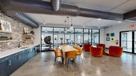 Located at 110 Field St in Newnan, GA, this single-floor amenity space blends industrial design with modern comfort across a generously sized open layout. Polished concrete floors, exposed structural ductwork, and soaring ceilings give the space a distinctive loft-like character throughout.

The fitness center is enclosed by sleek black steel-framed glass partitions, offering full visibility into the well-equipped workout area, which features cardio machines, weight racks, and ceiling fans. Natural light pours in through multi-pane windows, enhancing the airy feel.

Adjacent to the fitness area, a communal lounge and dining zone features a wooden dining table with tan leather chairs and plush burnt-orange velvet seating clusters. A kitchenette with blue-gray cabinetry and open shelving provides a functional refreshment station nearby.

A café-style nook lines an exposed brick accent wall adorned with a neon "This Must Be The Place" sign, with leather banquette booths and bistro tables creating an inviting social corner. The game room area centers on a ping-pong table and opens to a relaxed lounge with a blue sofa, mid-century credenza, and large windows overlooking the outdoors. Track lighting and pendant fixtures complete the well-layered illumination throughout this dynamic, multifunctional community space.