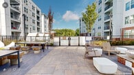 This expansive outdoor courtyard in Wake Forest, NC is a thoughtfully designed communal space that combines modern aesthetics with functional outdoor living. Enclosed by a contemporary multi-story building featuring a striking mix of brick and light gray siding, the courtyard offers a welcoming environment under open skies.

At the heart of the space, a wide stone-paved patio serves as the primary gathering area. A central fire pit lounge, surrounded by slatted wood Adirondack-style chairs and clean-lined white ottomans, provides an inviting spot for relaxation and socializing. Nearby, a long communal table paired with rustic wood block seating offers an ideal setting for outdoor dining or casual gatherings.

A manicured green lawn anchors one side of the courtyard, complemented by suspended hammocks that add a leisurely, resort-like touch. Mature trees and carefully curated plantings with gravel mulching borders line the perimeter, adding natural texture and greenery throughout the space.

The main building entrance is framed by a grand glass facade with floor-to-ceiling windows, flanked by upright evergreen shrubs and ornamental trees along a broad paved walkway. Perimeter fencing with a gated access point opens toward surrounding tree views. The overall ambiance is modern, open, and community-oriented, ideal for outdoor relaxation and social engagement.
