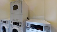 This single-floor laundry space is purpose-built to handle laundry needs with commercial-grade efficiency. The room is outfitted with a stacked washer and dryer unit alongside a freestanding Speed Queen Commercial Dryer, signaling a robust capacity suited for high-volume use. The machines feature digital control panels, offering precise settings for a variety of laundry cycles. The walls are finished in a clean, neutral tone, contributing to a bright and utilitarian atmosphere. The angled ceiling suggests the room may be tucked beneath a staircase or roofline, making practical use of an otherwise underutilized area. The layout is compact yet functional, with the equipment arranged to maximize the available floor space. The overall design prioritizes practicality, with durable, easy-to-clean surfaces throughout. This laundry space is well-suited for shared or high-demand use, offering dependable performance through its commercial appliances.