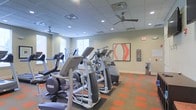 This fitness center at 1982 Route 70 East in Cherry Hill, New Jersey, offers a well-appointed workout space designed to support an active lifestyle. The facility is laid out on a single floor, providing an open and accessible environment for exercise. The room is equipped with a comprehensive array of modern cardio machines, including ellipticals and other aerobic equipment, arranged to maximize usability and floor space. Underfoot, a distinctive two-tone carpet in orange and neutral tones adds a dynamic visual element while providing comfort and durability. The ceiling is fitted with a suspended tile grid system that houses recessed lighting and ceiling fans, ensuring the space remains bright and well-ventilated throughout workouts. Natural light filters in through windows along one wall, further enhancing the open, energizing atmosphere. The neutral-toned walls are accented with contemporary artwork, including a bold abstract piece that adds a splash of color and personality to the space. A hand sanitizer or wipe station is also visible, reflecting a commitment to cleanliness and user well-being. Overall, the fitness center combines functional equipment, thoughtful design, and a welcoming ambiance to create an effective and comfortable exercise environment.