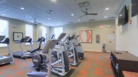 This fitness center at 1982 Route 70 East in Cherry Hill, New Jersey, offers a well-appointed workout space designed to support an active lifestyle. The facility is laid out on a single floor, providing an open and accessible environment for exercise. The room is equipped with a comprehensive array of modern cardio machines, including ellipticals and other aerobic equipment, arranged to maximize usability and floor space. Underfoot, a distinctive two-tone carpet in orange and neutral tones adds a dynamic visual element while providing comfort and durability. The ceiling is fitted with a suspended tile grid system that houses recessed lighting and ceiling fans, ensuring the space remains bright and well-ventilated throughout workouts. Natural light filters in through windows along one wall, further enhancing the open, energizing atmosphere. The neutral-toned walls are accented with contemporary artwork, including a bold abstract piece that adds a splash of color and personality to the space. A hand sanitizer or wipe station is also visible, reflecting a commitment to cleanliness and user well-being. Overall, the fitness center combines functional equipment, thoughtful design, and a welcoming ambiance to create an effective and comfortable exercise environment.