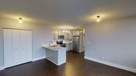 This single-floor space offers a thoughtfully designed open-concept layout that seamlessly connects the kitchen and living area, creating a bright and functional environment. The kitchen is outfitted with stainless steel appliances, including a refrigerator, electric range, and over-the-range microwave, all complemented by crisp white cabinetry that extends to the ceiling. A peninsula with an integrated sink and light countertops serves as both a functional workspace and a natural divider between the kitchen and the open living area. Overhead, a tasteful chandelier with multiple bulbs illuminates the kitchen zone, while a flush-mount ceiling light adds warmth to the adjacent hallway area. The rich, dark hardwood-style flooring runs throughout the space, providing a striking contrast against the light-toned walls and cabinetry. The clean lines, neutral color palette, and well-considered layout give the space a modern yet welcoming ambiance. With its open design and efficient use of space, this unit is well-suited for those who appreciate a contemporary and practical living environment.