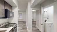This efficiently designed 2-bedroom, 1-bathroom unit makes smart use of its single-floor layout. The interior is finished with light gray luxury vinyl plank flooring throughout, complemented by crisp white walls, textured ceilings, and clean white baseboard trim that give the space a fresh, updated feel.

The kitchen is a standout feature, outfitted with dark gray shaker-style cabinetry accented by gold hardware, white subway tile backsplash, a stainless steel refrigerator, a gas range with over-the-range microwave, and a single-basin sink — all combining functionality with contemporary style.

The primary bedroom is the most spacious room in the unit, offering a window with blinds and a window-mounted air conditioning unit, providing natural light and climate control. The second bedroom mirrors a similar layout with its own window, baseboard heating, and ample wall space.

The bathroom has been renovated with white subway tile surrounding a full soaking tub, a modern square rain showerhead, and matte black fixtures, creating a clean, spa-like aesthetic. A central hallway connects the rooms, providing a logical and accessible flow throughout the unit. The overall ambiance is clean, modern, and move-in ready.