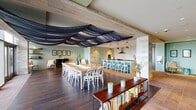 This thoughtfully designed single-floor space combines several distinct functional zones within a seamless open-plan layout. A dramatic navy fabric installation drapes across the exposed wood-beam ceiling, serving as a striking architectural focal point that visually connects the dining and kitchen areas. Wide-plank hardwood flooring grounds the dining zone, which accommodates a large communal table surrounded by white bentwood chairs, with floor-to-ceiling glass doors opening directly onto an outdoor terrace offering expansive views of the surrounding landscape.

The kitchen is a standout feature, defined by bold patterned tile walls, slate-toned cabinetry, warm yellow lower cabinets, and a generous white quartz island. A bar-height counter with teal cabinetry and pendant lighting in terracotta red provides additional seating and serves as a natural transition between the kitchen and dining areas.

A dedicated lounge zone features built-in, backlit bookshelves flanking a central television and media wall adorned with graphic wallpaper. Comfortable seating, a patterned ottoman, and layered rugs create an inviting, library-like atmosphere. An adjacent sitting area with sage-green walls, a mustard sofa, and curated artwork provides a quieter retreat. A low bookshelf console further emphasizes the space's literary character throughout.
