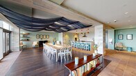 This thoughtfully designed single-floor space combines several distinct functional zones within a seamless open-plan layout. A dramatic navy fabric installation drapes across the exposed wood-beam ceiling, serving as a striking architectural focal point that visually connects the dining and kitchen areas. Wide-plank hardwood flooring grounds the dining zone, which accommodates a large communal table surrounded by white bentwood chairs, with floor-to-ceiling glass doors opening directly onto an outdoor terrace offering expansive views of the surrounding landscape.

The kitchen is a standout feature, defined by bold patterned tile walls, slate-toned cabinetry, warm yellow lower cabinets, and a generous white quartz island. A bar-height counter with teal cabinetry and pendant lighting in terracotta red provides additional seating and serves as a natural transition between the kitchen and dining areas.

A dedicated lounge zone features built-in, backlit bookshelves flanking a central television and media wall adorned with graphic wallpaper. Comfortable seating, a patterned ottoman, and layered rugs create an inviting, library-like atmosphere. An adjacent sitting area with sage-green walls, a mustard sofa, and curated artwork provides a quieter retreat. A low bookshelf console further emphasizes the space's literary character throughout.