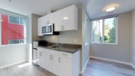This single-floor unit presents a clean, contemporary design throughout, making efficient use of its layout with a cohesive, modern aesthetic. The kitchenette is a standout feature, outfitted with glossy white flat-front cabinetry, dark countertops, a stainless steel microwave, a compact cooktop with integrated sink, and a stainless steel refrigerator — all finished with brushed metal hardware for a polished look. Light wood-tone flooring runs seamlessly throughout the space, lending warmth to the otherwise crisp, neutral palette of soft grey and white walls. The living area benefits greatly from a striking corner window installation that floods the space with natural light and offers pleasant views of surrounding greenery and neighboring structures, creating an airy, open feel. The bedroom area is bright and simply appointed, with a window that continues the theme of natural light and a wall-mounted heating unit for year-round comfort. The bathroom is thoughtfully designed with a tiled walk-in shower featuring classic white subway tile, a standard toilet, and wood-look flooring that ties back to the rest of the unit. A sliding door adds a space-conscious touch to the bathroom entry. Overall, the space offers a fresh, functional, and well-lit environment with consistent modern finishes throughout.