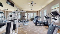 Located at Bristol Village in Midlothian, Virginia, this fitness center offers a comprehensive workout environment within a well-designed, single-floor layout. The space is thoughtfully appointed with warm neutral tones, taupe walls, and a patterned carpet flooring that adds texture underfoot. Elevated ceilings with dual ceiling fans promote airflow throughout, while recessed lighting ensures the room is consistently well-lit.

Large multi-pane windows line the walls on multiple sides, flooding the interior with natural light and providing pleasant views of the surrounding greenery. The abundance of natural light creates an open, energizing atmosphere ideal for exercise.

The fitness center is fully equipped to support a range of workout routines. Cardio equipment includes multiple treadmills, elliptical trainers, and a stationary cycle, all positioned to take advantage of the natural light and outdoor views. The strength training area features a multi-station cable machine, a weight bench with barbell rack, and a three-tier dumbbell rack stocked with a full range of free weights. Wall-mounted mirrors expand the sense of space and allow users to monitor their form during workouts. Wall-mounted televisions provide entertainment options. The space also includes sanitation stations and clearly defined entry doors, contributing to a clean and organized environment.