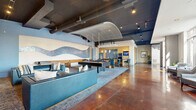 Located at 951 Fell St in Baltimore's vibrant Fells Point neighborhood, this contemporary amenity lounge is a well-appointed communal space designed for both relaxation and entertainment. The open-plan layout spans a generous single floor, seamlessly connecting multiple activity zones beneath dramatic exposed ceilings with dark-painted ductwork and beam details that lend the space an industrial-modern character.

Polished concrete floors in a rich, warm amber tone anchor the open areas, transitioning to plush, patterned area rugs in the carpeted lounge and game zones. A dedicated billiards area serves as a social centerpiece, featuring a full-size pool table beneath an elegant arched ceiling detail, flanked by teal upholstered seating and accented by a striking wave-patterned mosaic tile wall. Adjacent to this, a media lounge offers multiple sofas and a large flat-screen entertainment setup, with a shuffleboard table nearby for additional recreation.

The full kitchen and bar area, outfitted with warm wood cabinetry, a mosaic tile backsplash, stainless steel appliances, and pendant lighting, provides a functional space for hosting and entertaining. Floor-to-ceiling windows along one perimeter flood the space with natural light and offer direct views of the outdoor pool courtyard, where a bright seating area is visible just beyond. Curated artwork and greenery add finishing touches throughout, creating an inviting and polished atmosphere.