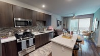 This well-appointed single-floor residence seamlessly blends modern design with comfortable living. The open-concept layout connects the kitchen and living area, creating an inviting and functional space. The kitchen is outfitted with stainless steel appliances, including a gas range, built-in microwave, and dishwasher, complemented by rich dark wood cabinetry and a mosaic tile backsplash. A generous white waterfall-edge peninsula provides additional prep space and doubles as a breakfast bar with seating. Dark hardwood-style flooring runs throughout, grounding the space with warmth and continuity. The living area is bright and welcoming, highlighted by a bold slate-blue accent wall that adds depth and character. Large sliding glass doors flood the room with natural light and open directly onto a private balcony, where outdoor seating offers a seamless transition between indoor and outdoor living. A ceiling fan keeps the space comfortable year-round. The overall ambiance is contemporary and curated, with thoughtful design choices — from the patterned area rug to the artistic wall décor — that create a cohesive and stylish atmosphere throughout.