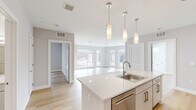This contemporary single-floor residence showcases a clean, modern design highlighted by a neutral palette of soft grays and crisp whites throughout. The heart of the home is the open-concept kitchen and living area, where a generously sized white quartz island with an undermount sink serves as a central hub. Three sleek pendant lights hang above the island, providing both task lighting and visual interest. The kitchen is fully equipped with stainless steel appliances, including a refrigerator, electric range, built-in microwave, and dishwasher, all complemented by white cabinetry with brushed nickel hardware and a subtle tile backsplash. Light-toned hardwood flooring flows seamlessly throughout the entire unit, enhancing the sense of openness and continuity. The living area benefits from an impressive bank of floor-to-ceiling windows on two walls, flooding the space with natural light and offering views of the surrounding streetscape. The bedrooms are well-proportioned, each featuring large windows that bring in additional daylight. The bathroom is finished with marble-style tile flooring, clean white walls, a tiled shower surround, and brushed metal grab bars. Overall, the unit presents a bright, airy, and thoughtfully designed living environment with a consistently modern aesthetic.