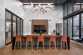 This sophisticated single-floor amenity lounge is defined by its striking architectural design and carefully curated interiors. Soaring double-height ceilings create an impressive sense of volume, while expansive floor-to-ceiling glass walls flood the space with abundant natural light and offer unobstructed views of an adjacent outdoor terrace. The herringbone-patterned hardwood flooring runs throughout, lending warmth and continuity to the space.

The main lounge area is anchored by a large, comfortable sectional sofa paired with a marble-topped coffee table, complemented by an eclectic arrangement of accent chairs in rich navy and orange tones. A sculptural, multicolored glass-globe chandelier serves as a bold artistic focal point overhead. A built-in media wall with wood paneling and integrated cabinetry provides a mounted flat-screen TV, creating a dedicated entertainment zone. An elongated counter-height work surface with vibrant orange bar seating offers a functional co-working or dining area.

A compact kitchenette, tucked discreetly behind a floor-length curtain, features dark cabinetry, a light quartz countertop, an under-counter refrigerator, and floating wood shelves with under-shelf lighting. Black-framed glass partition walls and sliding glass doors divide zones while maintaining visual openness. Additional corridors with textured wallcoverings and wood-railed half-walls connect supporting areas, completing this well-appointed and design-forward communal space.