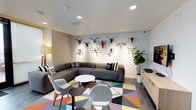 This single-floor space makes a striking first impression with its creative, contemporary design. The focal point of the lounge area is an eye-catching feature wall adorned with a collection of colorful, sculptural animal head mounts — rendered in vivid hues of red, blue, yellow, and green — lending the space a playful, artistic character that sets it apart from conventional interiors.

The seating area is anchored by a generous L-shaped sectional sofa in charcoal gray, complemented by a pair of sleek, modern accent chairs in light gray. A round white coffee table sits at the center, positioned atop a bold geometric rug in a lively mix of teal, yellow, orange, purple, and gray tones that adds energy and warmth to the floor.

Recessed lighting in the white ceiling provides clean, even illumination throughout, enhancing the bright and open atmosphere of the room. A tall fiddle-leaf fig plant in the corner introduces a natural element, softening the space alongside a light wood credenza that offers functional storage. Decorative throw pillows with playful graphics add personality to the seating arrangement. Overall, the space balances a fun, expressive aesthetic with practical, comfortable furnishings — making it well-suited for relaxed socializing or informal gatherings.