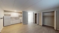 This single-floor unit offers a straightforward, functional layout designed for efficient everyday living. The open main living area is finished with light wood-tone laminate flooring and neutral gray walls, creating a clean and understated atmosphere. A baseboard heating system runs along the perimeter, ensuring consistent warmth throughout the space.

The kitchen is positioned along one wall of the main area and is equipped with white raised-panel cabinetry spanning both upper and lower sections, providing ample storage. A double-basin stainless steel sink, a light-toned laminate countertop, a white electric coil range, and a full-size refrigerator complete the kitchen's practical setup.

The bedroom features darker wood-tone laminate flooring and includes two side-by-side bifold closets, offering generous built-in storage. A window with blinds allows for natural light, and a wall-mounted air conditioning unit provides climate control.

The bathroom is bright and clean, finished in white throughout. It includes a full bathtub with a shower curtain rod, a toilet with a grab bar for accessibility, and light laminate flooring consistent with the rest of the unit.

A thermostat is conveniently located near the bathroom entrance, and the overall design prioritizes clean lines, low-maintenance finishes, and practical functionality throughout every area of the space.