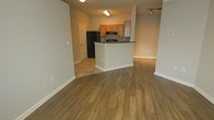 This single-floor space is designed with an open-concept layout that promotes a sense of spaciousness and functional flow. The living area features warm, medium-toned hardwood-style flooring that extends throughout, adding a cohesive and inviting aesthetic to the space. Neutral gray walls provide a clean, versatile backdrop suitable for a variety of interior styles.

The kitchen is positioned centrally within the open floor plan and is defined by a peninsula with a dark countertop, offering both additional workspace and a natural visual divider between the kitchen and living areas. Honey-toned wood cabinetry lines the kitchen walls, providing ample storage, while black appliances — including a refrigerator and range with overhead microwave — offer a contrasting, modern touch. Track lighting in the adjoining area and flush ceiling fixtures throughout ensure adequate illumination across the space.

A doorway visible near the kitchen leads to an additional room, suggesting further functional areas beyond the main open space. The flooring transitions subtly near the kitchen, where tile or a lighter material meets the hardwood-style planks of the living area. Overall, the space presents a practical, well-organized layout with a warm and neutral ambiance.