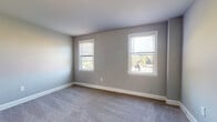 This single-floor residence offers a well-organized layout with multiple rooms finished in a consistent neutral gray palette, creating a calm and cohesive atmosphere throughout. The bedrooms are carpeted and generously sized, each featuring multiple windows with slatted blinds that invite natural light and offer views of surrounding greenery. White baseboards provide a clean contrast against the gray walls. Several bedrooms are equipped with wall-mounted mini-split units for individual climate control, and at least one room includes a ceiling fan for additional air circulation. The kitchen is a compact and functional space outfitted with white cabinetry, a coil-burner electric range, a single-basin sink, and laminate countertops. A window above the kitchen area brings in natural light and views of the outdoors. The bathroom features a clean, minimalist design with white subway tile lining the tub surround, wood-look vinyl flooring, a pedestal-style vanity, and a window for natural ventilation. The floor plan, as seen in the dollhouse view, reveals a practical and efficient arrangement of rooms connected by a central corridor, making the space easy to navigate and functional for everyday living.