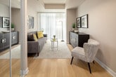 Located at 111 Dundas St W in the heart of Toronto, this single-floor unit offers a well-organized layout designed for comfortable urban living. An entry hallway, accented by a wood-framed mirror, connects the various areas of the space and transitions from tile flooring to light hardwood as you move deeper into the unit.

The living area is bright and welcoming, featuring large floor-to-ceiling windows dressed with vertical blinds that allow generous natural light. The space accommodates a seating arrangement anchored by a tufted grey sofa, complementary accent pillows in yellow and black, and a bold large-scale black-and-white abstract artwork on the wall, lending the room a contemporary character.

The kitchen is efficiently designed with dark wood-grain cabinetry extending to the ceiling, providing ample storage. Black appliances — including a range, dishwasher, and refrigerator — are paired with light countertops and a stainless steel sink. A breakfast bar with bar seating extends from the counter, offering a casual dining option alongside a dedicated dining area nearby.

The bathroom features a full soaking tub with a shower, finished with white and tan accent tile surround, neutral tile flooring, and decorative coastal-themed wall art, creating a clean and relaxed atmosphere throughout.