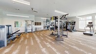 Located at 4001 Reedybrook Crossing in Apex, NC, this comprehensive fitness center offers a generously sized, single-floor layout thoughtfully divided into distinct workout zones. The main strength and cardio area features patterned, textured carpeting and is equipped with multi-station cable machines, a functional training rig with TRX suspension straps, a punching bag, free weights, kettlebells, a dumbbell rack, and a selection of cardio machines including treadmills and ellipticals. Full-length mirrors along the walls amplify the sense of space, while ceiling fans and linear pendant lighting maintain a bright, comfortable environment. Large windows throughout flood the area with abundant natural light and offer views of the surrounding neighborhood. A dedicated cycling studio showcases light wood-look flooring, a row of spin bikes, and a rack of stability balls and resistance accessories. An adjoining multi-purpose room features the same wood-look flooring, a soft teal accent wall, wall-mounted display screen, and built-in cabinetry — ideal for classes or training sessions. A bold black-and-white fitness-themed photo mural adds an energetic, motivational accent to the strength zone. The overall design is modern and clean, creating an inviting and functional environment for a wide range of fitness activities.