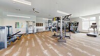 Located at 4001 Reedybrook Crossing in Apex, NC, this comprehensive fitness center offers a generously sized, single-floor layout thoughtfully divided into distinct workout zones. The main strength and cardio area features patterned, textured carpeting and is equipped with multi-station cable machines, a functional training rig with TRX suspension straps, a punching bag, free weights, kettlebells, a dumbbell rack, and a selection of cardio machines including treadmills and ellipticals. Full-length mirrors along the walls amplify the sense of space, while ceiling fans and linear pendant lighting maintain a bright, comfortable environment. Large windows throughout flood the area with abundant natural light and offer views of the surrounding neighborhood. A dedicated cycling studio showcases light wood-look flooring, a row of spin bikes, and a rack of stability balls and resistance accessories. An adjoining multi-purpose room features the same wood-look flooring, a soft teal accent wall, wall-mounted display screen, and built-in cabinetry — ideal for classes or training sessions. A bold black-and-white fitness-themed photo mural adds an energetic, motivational accent to the strength zone. The overall design is modern and clean, creating an inviting and functional environment for a wide range of fitness activities.