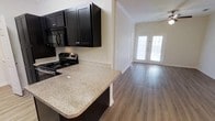 This single-story space showcases a thoughtfully designed open-concept layout that seamlessly connects the kitchen and living area. The kitchen is outfitted with rich, dark espresso cabinetry that provides a striking contrast against the light granite countertops. Stainless steel appliances, including a gas range and built-in microwave, add a modern, functional touch. A peninsula-style counter extends into the space, creating a natural division between the kitchen and the adjacent living area while offering additional workspace or casual seating potential.

Crown molding along the ceiling adds an elegant architectural detail, lending a refined character to the interior. The warm, medium-toned hardwood-style flooring flows continuously throughout both areas, contributing to a cohesive and polished aesthetic. The living area benefits from natural light streaming through a set of double French doors, which serve as a focal point at the far end of the room. The neutral wall tones complement both the dark cabinetry and flooring, creating a balanced and inviting ambiance throughout the space.