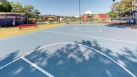 Located at 19 Landmark Dr in Columbia, South Carolina, this outdoor recreational sports facility features a spacious, blue-surfaced multi-use court designed for basketball and additional court sports. The court is clearly marked with crisp white boundary and key lines, and is equipped with a regulation basketball hoop and backboard mounted on a sturdy pole. The entire area is enclosed by green chain-link fencing, providing a secure and defined play space.

The court is set within a lush, tree-lined environment, with mature trees casting dappled shade across the surface — offering natural relief from the South Carolina sun. The surrounding grounds reveal a vibrant community setting, with a colorful playground area visible adjacent to the court, along with seating and shaded picnic areas equipped with yellow umbrellas nearby.

Brick residential buildings frame portions of the perimeter, alongside a blue utility or community building. The natural landscaping, including shrubs and grassy areas, integrates the court smoothly into its surroundings. The overall ambiance is active and community-oriented, making this a well-positioned recreational amenity suited for both organized play and casual outdoor activity throughout the year.