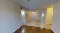 This single-floor space offers a straightforward, functional layout with a notable transition between two distinct flooring types — rich hardwood floors in the entry area giving way to light-toned ceramic tile throughout the main living space. The open floor plan creates a sense of continuity between the living and dining areas, allowing for flexible furniture arrangements. A ceiling fan is centrally positioned in the main living area, providing both air circulation and ambient lighting. The textured popcorn ceiling and neutral cream-toned walls provide a clean, understated backdrop throughout. A partially visible kitchen is situated to the right, equipped with stainless steel appliances including a range, and features light cabinetry and countertops. The space benefits from interior lighting, with a wall-mounted fixture visible near what appears to be a closet or utility area. White baseboards frame the rooms cleanly, and an electrical outlet is conveniently placed along the lower wall. Overall, the space presents a practical and livable layout suited for everyday use, with durable finishes and a neutral palette that offers versatility for personalization.