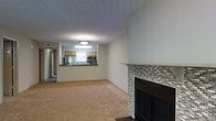 This single-floor residence offers a welcoming and functional layout designed for comfortable everyday living. The main living area is anchored by a standout wood-framed fireplace adorned with a detailed mosaic tile surround and a matching tiled mantel, adding a stylish focal point to the room. Plush wall-to-wall carpeting in a warm neutral tone runs throughout the living space, contributing to a cozy and inviting atmosphere. The textured ceiling adds a classic touch to the interior. The living area flows seamlessly into the kitchen, which is positioned at a raised bar-style counter that serves as both a functional workspace and a casual dining divider. The kitchen is equipped with stainless steel appliances, including a refrigerator and microwave, and features warm wood-toned cabinetry paired with dark countertops. A window above the kitchen sink allows natural light to brighten the cooking space. A ceiling-mounted light fixture provides additional illumination. The open-concept design between the living and kitchen areas creates a connected, airy feel throughout the main living space, making it well-suited for both daily routines and casual entertaining.
