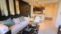 This single-floor residence showcases an open-concept layout that integrates the living, dining, and kitchen areas into one fluid, well-appointed space. The living area is anchored by a crisp white sofa dressed with patterned and solid accent pillows, positioned atop a black-and-white geometric area rug. A dramatic black accent wall serves as a focal point, adorned with a decorative starburst clock and a tall mirror that enhances the sense of depth and light.

Adjacent to the living area, a glass-top dining table surrounded by elegant white French-style chairs creates an intimate setting that bridges the living space and kitchen. The kitchen features dark espresso cabinetry, stainless steel appliances, and a peninsula countertop with warm stone-toned surfaces, offering both functionality and visual appeal. Recessed lighting throughout ensures the space is well-lit, complementing the neutral carpeted flooring that runs through the main living areas. The overall ambiance balances bold, contrasting design elements with clean lines, achieving a polished and contemporary aesthetic.