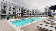 Located at 203 3rd Ave in Asbury Park, NJ, this beautifully designed courtyard amenity space serves as a central outdoor hub surrounded by a modern multi-story residential building with contemporary gray siding, large windows, and private balconies. The courtyard is thoughtfully organized into several distinct zones, each catering to a different aspect of outdoor living. A large, sparkling resort-style swimming pool anchors one end of the space, lined with sleek lounge chairs and navy blue umbrellas that provide shade along a wide pool deck. Adjacent to the pool, elevated decking areas offer additional lounging with modern outdoor furniture and greenery. Lush artificial turf lawns are interspersed throughout the courtyard, providing vibrant color and comfortable gathering areas. An outdoor entertainment zone features a large flat-screen TV, a bar-height counter, and a grill station, making it ideal for social gatherings. Intimate seating vignettes are defined by tall boxwood hedges, offering semi-private spaces for dining and conversation. Raised planters with seasonal plantings add natural texture throughout. Wide walkways and accessible ramps connect all areas seamlessly, while the surrounding building's multi-story facade creates a sense of enclosure and community within this well-appointed outdoor retreat.