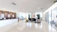 This contemporary professional space is thoughtfully designed to make a strong first impression. The open reception area anchors the space with a sleek, dark-wood reception desk that provides a warm contrast against the predominantly white and neutral palette throughout. Polished light stone flooring extends across the entire floor, lending an air of sophistication and cleanliness.

The seating lounge area is furnished with modern sofas and a distinctive yellow accent chair, arranged around a glass coffee table atop a patterned area rug, creating an inviting and stylish environment for guests and visitors. Vibrant potted bamboo and leafy plants are strategically placed throughout, introducing a refreshing organic element that softens the clean lines of the interior.

Glass-partitioned private offices or meeting rooms line the space, offering both transparency and acoustic separation while maintaining the open, airy feel of the overall layout. Recessed ceiling lighting combined with large floor-to-ceiling windows allows natural light to flood the interior, providing views of lush greenery outside. A colorful framed artwork piece adorns one wall, adding a creative focal point. The overall ambiance is professional, modern, and welcoming.