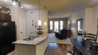 This single-floor residence offers an inviting open-concept design that seamlessly connects the kitchen and living areas, creating a cohesive and functional living environment. The kitchen is outfitted with a curved granite-style countertop peninsula that doubles as a breakfast bar, a double-basin stainless steel sink, and stylish pendant lighting that hangs above the counter, casting a warm glow over the space. Warm-toned walls complement the rich hardwood flooring that flows through the main living areas, adding depth and character to the interior.

The living room is generously sized, anchored by a cozy seating arrangement set atop a plush area rug. A ceiling fan overhead provides comfort, while a set of large double French doors floods the space with natural light and offers a view to the outdoors. A TV and floor lamp complete the living area, making it a comfortable and well-lit retreat. Neutral wall tones and clean architectural lines give the space a timeless, welcoming ambiance. Recessed lighting and ceiling-mounted fixtures provide additional illumination throughout, ensuring a bright and comfortable atmosphere at any time of day.