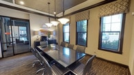 This single-floor conference room is a well-appointed meeting space designed for professional gatherings. At its center sits a large, rectangular glass-top table surrounded by ergonomic rolling chairs upholstered in gray fabric, providing comfortable seating for multiple attendees. Three pendant lights with frosted glass shades hang in a row above the table, casting warm, focused illumination throughout the room.

The interior is finished with dark wood crown molding that frames the textured ceiling and contrasts elegantly with the neutral beige walls. A patterned carpet featuring an abstract wave design in warm gold and brown tones adds visual interest underfoot. Along one wall, a dark credenza houses office equipment, including a printer, and is accented with decorative accessories and greenery.

Multiple windows dressed with geometric-patterned Roman shades line the exterior wall, allowing natural light to filter in while offering views of outdoor greenery. Glass-paneled doors provide transparency into the adjacent corridor, which connects directly to a fitness center visible through floor-to-ceiling glass partitions. A decorative circular metal wall accent and framed artwork contribute to the room's refined, contemporary aesthetic, making it a functional yet visually engaging professional environment.