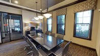 This single-floor conference room is a well-appointed meeting space designed for professional gatherings. At its center sits a large, rectangular glass-top table surrounded by ergonomic rolling chairs upholstered in gray fabric, providing comfortable seating for multiple attendees. Three pendant lights with frosted glass shades hang in a row above the table, casting warm, focused illumination throughout the room.

The interior is finished with dark wood crown molding that frames the textured ceiling and contrasts elegantly with the neutral beige walls. A patterned carpet featuring an abstract wave design in warm gold and brown tones adds visual interest underfoot. Along one wall, a dark credenza houses office equipment, including a printer, and is accented with decorative accessories and greenery.

Multiple windows dressed with geometric-patterned Roman shades line the exterior wall, allowing natural light to filter in while offering views of outdoor greenery. Glass-paneled doors provide transparency into the adjacent corridor, which connects directly to a fitness center visible through floor-to-ceiling glass partitions. A decorative circular metal wall accent and framed artwork contribute to the room's refined, contemporary aesthetic, making it a functional yet visually engaging professional environment.