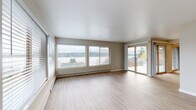This single-floor space is designed with a clean, neutral aesthetic throughout, featuring light wood-tone flooring in the main living areas and kitchen, complemented by soft carpet in the bedrooms. The living room is a standout area, offering generous proportions and abundant natural light through a wide bank of windows fitted with horizontal blinds. A sliding glass door connects the living space directly to an outdoor area, framing picturesque views of the surrounding landscape and water in the distance, creating a bright and airy atmosphere.

The kitchen is well-equipped with light wood cabinetry extending to the ceiling for ample storage, light-toned laminate countertops, and a full suite of appliances including a stainless gas range, dishwasher, and refrigerator. A flush ceiling light fixture provides practical illumination over the workspace.

The bedrooms are comfortably sized, each featuring plush carpet, ceiling fans, and sliding-door closets with built-in rods and shelving. Windows allow natural light to filter in. A hallway connects the rooms efficiently, with a sliding storage closet conveniently accessible along the corridor.

The bathroom includes a light stone-toned countertop with an undermount sink, wood-look flooring, and a glass-door shower enclosure, maintaining the space's consistent neutral and modern palette throughout.