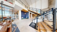 Located at 1250 Hitchings Way in Durham, NC, the Nixie Clubhouse is a generously scaled community facility that showcases contemporary architectural design across a single sprawling floor. The entrance lobby immediately impresses with soaring double-height ceilings and expansive floor-to-ceiling glass walls that flood the space with natural light and offer clear views of the surrounding streetscape. Large-format light stone tile flooring runs throughout, lending a clean, cohesive aesthetic.

A centerpiece floating staircase with warm wood treads, black steel framing, and glass railings serves as a dramatic architectural focal point. The main lounge area features an artful arrangement of seating, including leather sofas, accent chairs, and curated area rugs, anchored by a bold stone fireplace surround with built-in cabinetry and a mounted display screen. A wood-paneled tray ceiling with oversized ring pendant lighting defines a social lounge and dining zone with ample seating and access to a wet bar area.

Additional highlights include a reception desk backed by a striking geometric wood slat accent wall, glass-partitioned private office or conference spaces, and intimate lounge alcoves with tufted blue seating and pendant lighting. The overall ambiance is sophisticated yet welcoming, blending modern materials with warm wood tones throughout.